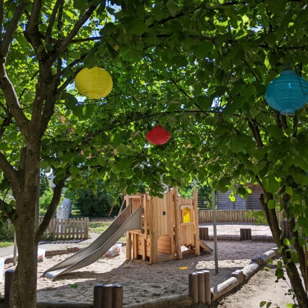 Ein hölzerner Spielplatz auf Sanduntergrund unter grünen Bäumen.